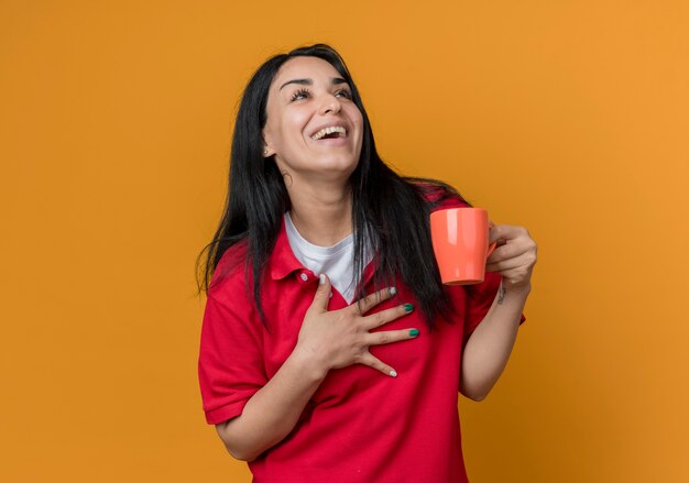 Alegre joven morena caucásica vestida con camisa roja pone la mano en el pecho y sostiene la taza mirando al lado aislado en la pared naranja