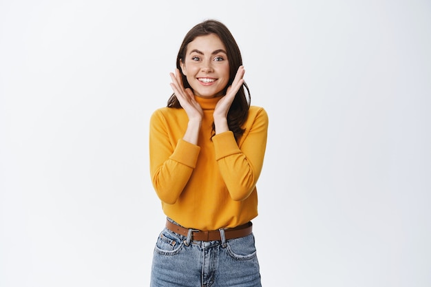 Alegre joven morena regocijándose del regalo sorpresa, sonriendo y mirando emocionado, de pie en suéter amarillo y jeans contra la pared blanca