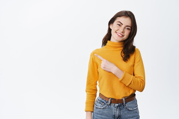 Alegre joven morena sonriendo señalando con el dedo a la izquierda en el logotipo que muestra espacio de copia para su anuncio de pie en suéter amarillo sobre fondo blanco.