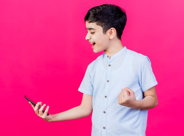 Alegre joven muchacho caucásico sosteniendo mirando el teléfono móvil haciendo sí gesto aislado sobre fondo carmesí