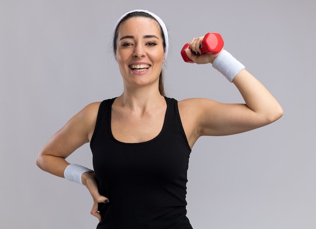 Alegre joven mujer bastante deportiva con diadema y muñequeras mirando al frente manteniendo la mano en la cintura levantando mancuernas aislado en la pared blanca