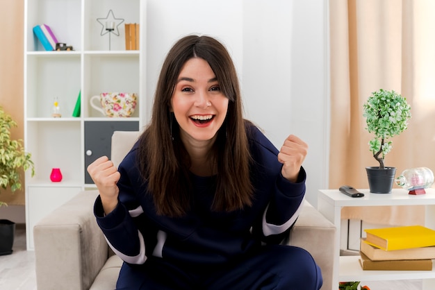 Alegre joven mujer bonita caucásica sentada en un sillón en la sala de estar diseñada mirando haciendo gesto de sí