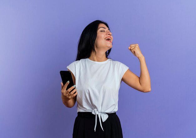 Alegre joven mujer caucásica mantiene el puño y sostiene el teléfono mirando a un lado