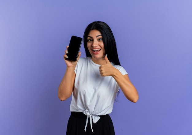 Alegre joven mujer caucásica sostiene el teléfono y los pulgares para arriba