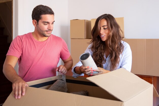 Alegre joven y mujer moviendo y desempacando cosas, abriendo la caja de cartón y sacando el objeto