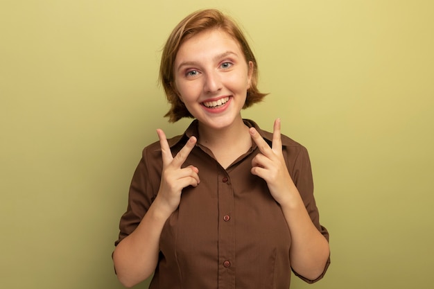 Alegre joven mujer rubia mirando al frente haciendo el signo de la paz aislado en la pared verde oliva con espacio de copia