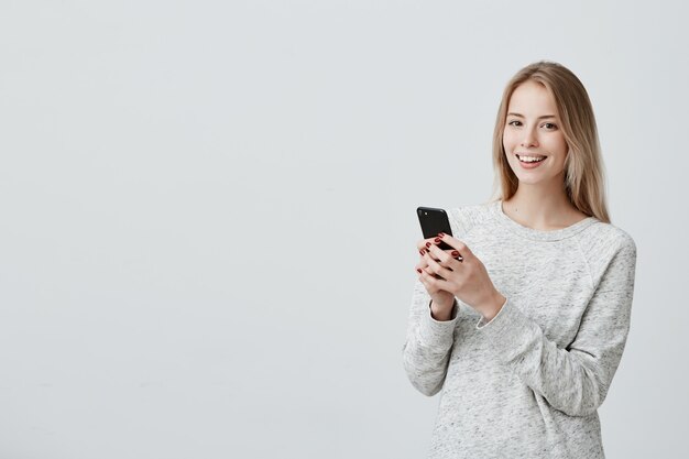 Alegre joven mujer rubia de pelo rubio con linda sonrisa posando en interiores, utilizando el teléfono celular, revisando el suministro de noticias en sus cuentas de redes sociales. Mujer bonita navegando por internet en el móvil