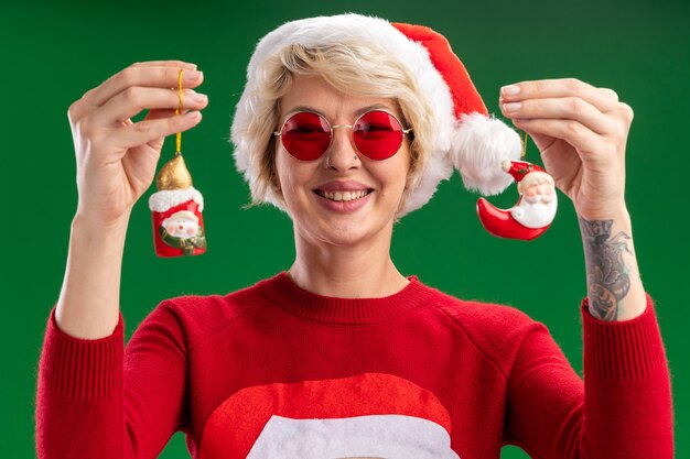 alegre joven mujer rubia con sombrero de navidad y santa claus suéter de navidad con gafas mirando mostrando muñeco de nieve y santa claus adornos navideños aislados en la pared verde