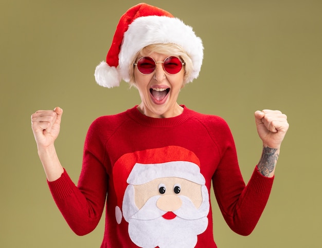 Foto gratuita alegre joven mujer rubia con sombrero de navidad y suéter de navidad de santa claus con gafas mirando haciendo gesto de sí aislado en la pared verde oliva