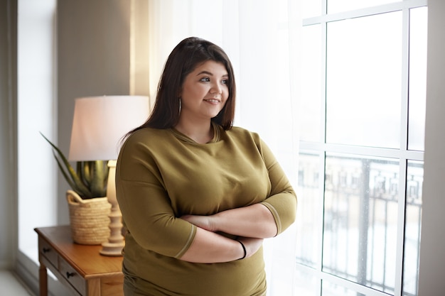 Foto gratuita alegre joven mujer con sobrepeso de talla grande con cabello negro y mejillas regordetas cruzando los brazos sobre su pecho y sonriendo felizmente, de pie junto a la ventana, mirando hacia afuera, contemplando la hermosa vista de la mañana