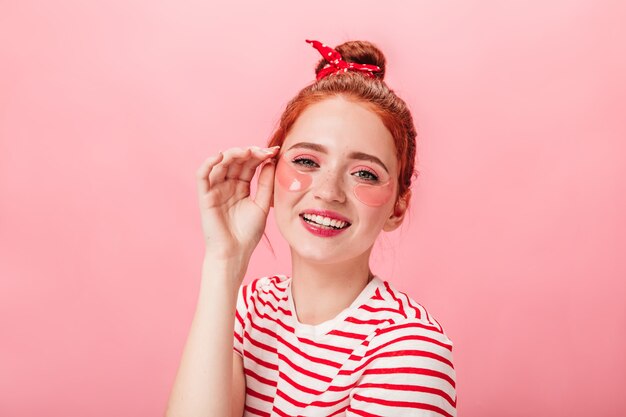 Alegre joven con parches en los ojos mirando a la cámara. Foto de estudio de chica jengibre emocionada haciendo tratamiento de cuidado de la piel sobre fondo rosa.