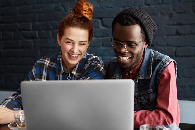 Alegre joven pareja interracial disfrutando de una conexión inalámbrica a Internet de alta velocidad en la cafetería, comprando en línea o viendo series en una computadora portátil genérica, mirando la pantalla con sonrisas felices y alegres