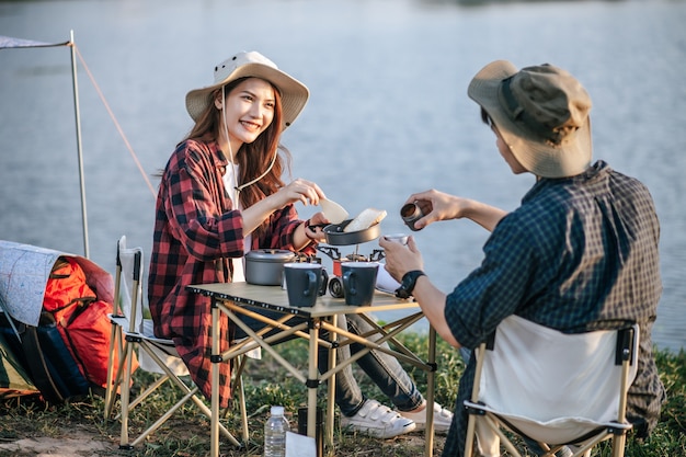 Alegre joven pareja de mochileros con sombrero de trekking sentado cerca del lago con café y desayuno y haciendo molinillo de café recién hecho mientras acampa en vacaciones de verano