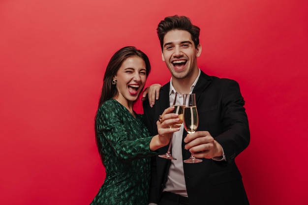 Foto gratuita alegre joven pareja de personas en trajes de vacaciones, sonriendo, sosteniendo copas con champán y mirando al frente aislado en la pared roja