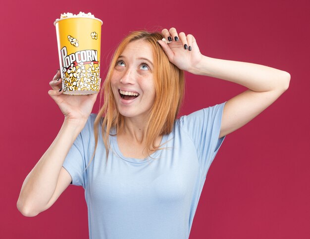 Alegre joven pelirroja pelirroja con pecas sosteniendo un cubo de palomitas de maíz y mirando hacia arriba