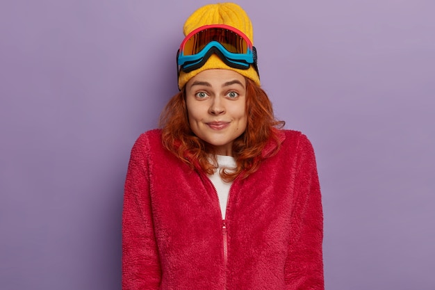 Foto gratuita alegre joven pelirroja viste ropa de abrigo, gafas de esquí en la cabeza, posa sobre fondo púrpura.