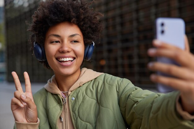 Alegre joven de pelo rizado hace selfie muestra el signo de la paz sonríe ampliamente utiliza auriculares inalámbricos