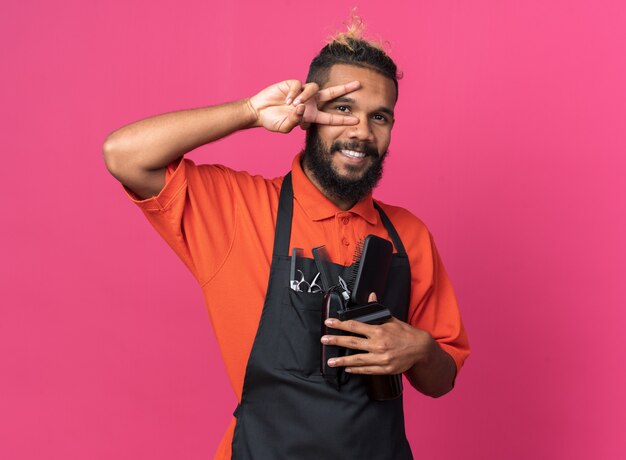 Alegre joven peluquero masculino afroamericano vistiendo uniforme sosteniendo herramientas de peluquero que muestra el símbolo de signo v cerca del ojo