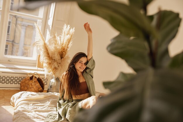 Alegre joven de piel clara sonriendo mirando a la cámara mientras se sienta en el suelo en una habitación cómoda Mujer morena con el pelo largo viste ropa informal Concepto de descanso y recuperación