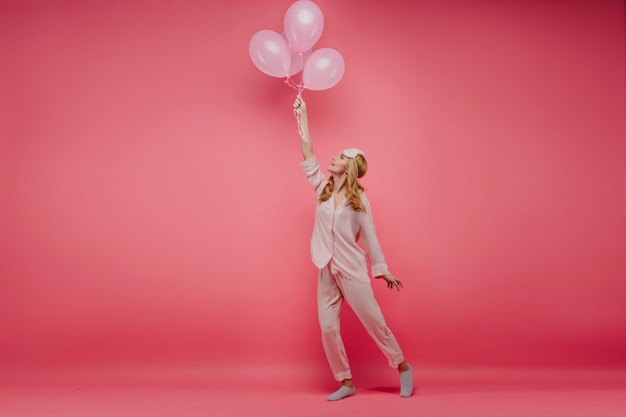 Alegre joven en pijama de seda bailando con globos de fiesta. Foto de cuerpo entero de la espectacular cumpleañera en traje de noche divertido posando en la pared rosa.