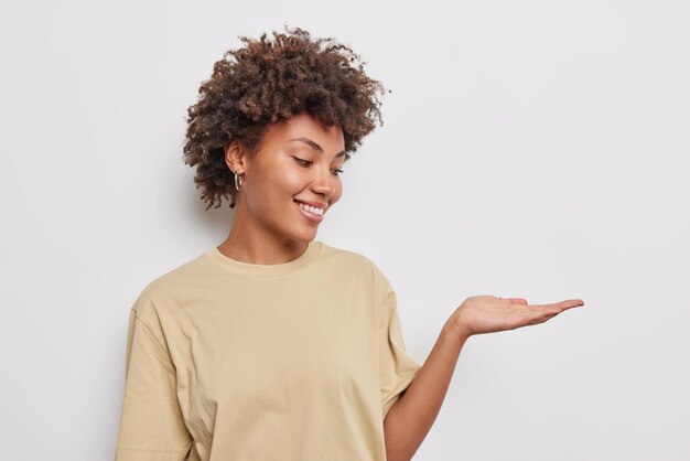 Alegre joven positiva de pelo rizado finge sostener algo mantiene la palma levantada vestida con una camiseta beige casual aislada sobre fondo blanco, espacio de copia en blanco para su contenido publicitario