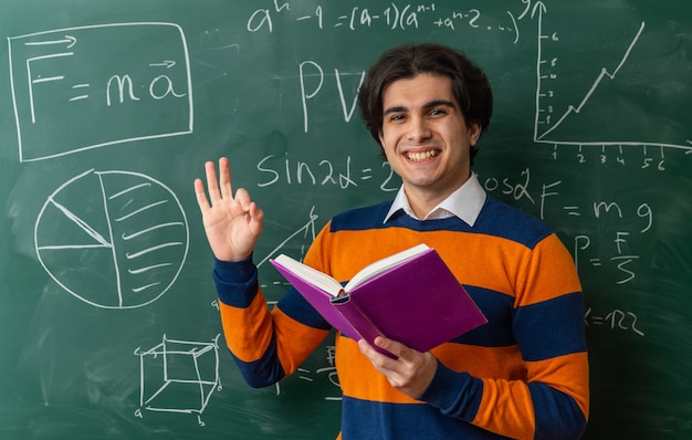 Alegre joven profesor de geometría de pie delante de la pizarra en el aula sosteniendo el libro mirando al frente haciendo bien firmar