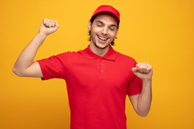 alegre joven repartidor con uniforme y gorra manteniendo los puños en el aire con los ojos cerrados aislado sobre fondo amarillo