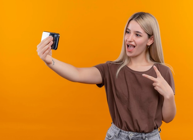 Alegre joven rubia en aparatos dentales haciendo el signo de la paz y tomando selfie en un espacio naranja aislado