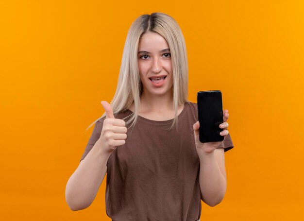Alegre joven rubia en aparatos dentales sosteniendo el teléfono móvil mostrando el pulgar hacia arriba en el espacio naranja aislado con espacio de copia