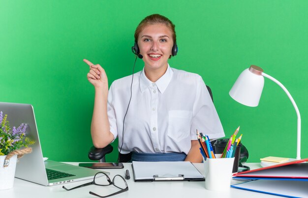 Alegre joven rubia centro de llamadas chica con auriculares sentado en el escritorio con herramientas de trabajo mirando a la cámara apuntando al lado aislado en la pared verde