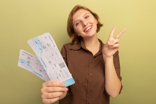 Alegre joven rubia estirando los billetes de avión haciendo el signo de la paz