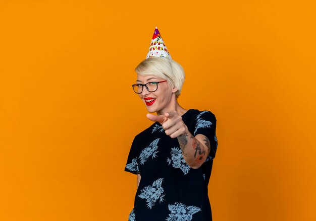 Alegre joven rubia fiestera con gafas y gorra de cumpleaños mirando y apuntando a la cámara aislada sobre fondo naranja con espacio de copia