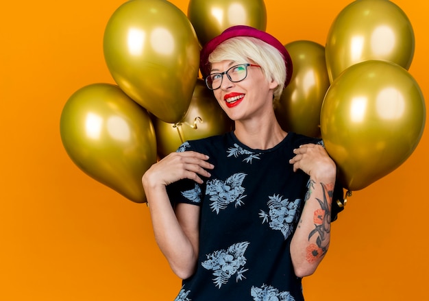 Foto gratuita alegre joven rubia fiestera con gorro de fiesta y gafas de pie delante de globos mirando al lado tocando los hombros aislados sobre fondo naranja