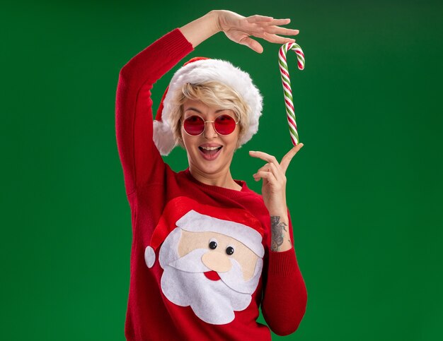 Alegre joven rubia con sombrero de navidad y suéter de navidad de santa claus con gafas mirando sosteniendo bastón de caramelo de navidad cerca de la cabeza aislada en la pared verde con espacio de copia