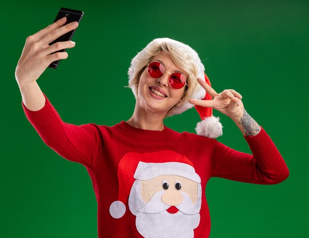 Alegre joven rubia vistiendo gorro de navidad y suéter de navidad de santa claus con gafas haciendo el signo de la paz tomando selfie aislado sobre fondo verde