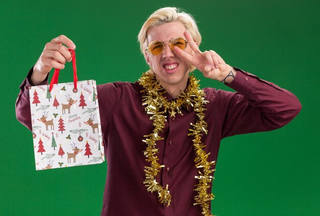 Alegre joven rubio con gafas con guirnalda de oropel alrededor del cuello sosteniendo una bolsa de regalo de Navidad haciendo el signo de la paz aislado en la pared verde