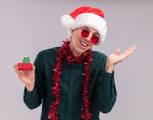 Alegre joven rubio con gorro de Papá Noel y gafas con guirnalda de oropel alrededor del cuello sosteniendo el juguete del árbol de Navidad con fecha mirando a cámara mostrando la mano vacía aislada sobre fondo blanco