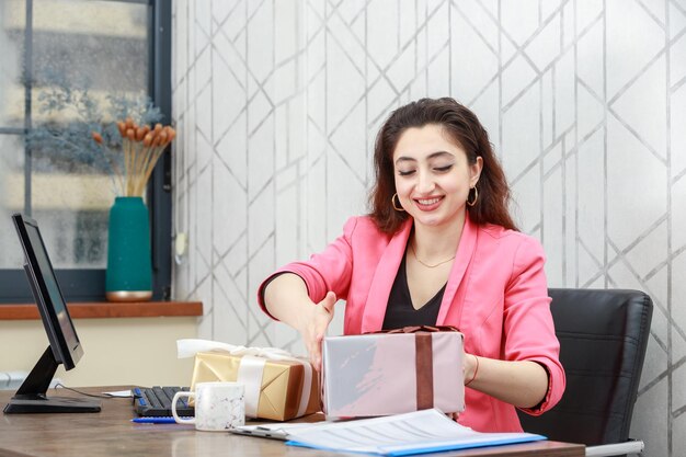 La alegre joven sentada detrás del escritorio y abriendo su caja de regalo Foto de alta calidad