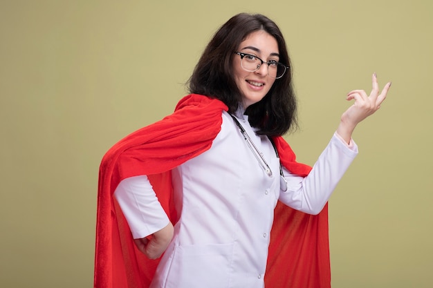 Alegre joven superhéroe caucásica en capa roja con uniforme médico y estetoscopio con gafas de pie en vista de perfil manteniendo las manos en el aire aislado en la pared verde oliva