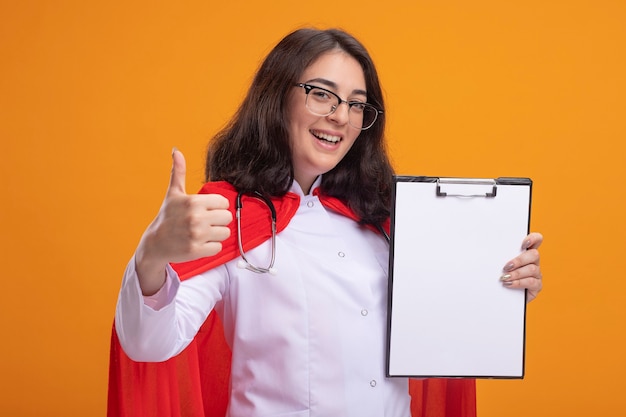 Alegre joven superhéroe caucásica en capa roja vistiendo uniforme médico y estetoscopio con gafas mostrando el portapapeles a la cámara mostrando el pulgar hacia arriba