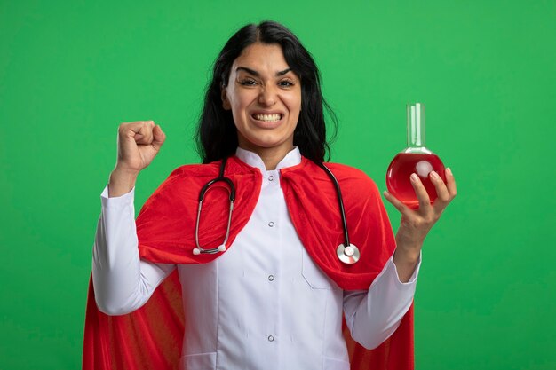 Alegre joven superhéroe vestida con bata médica con estetoscopio sosteniendo una botella de vidrio de química llena de líquido rojo que muestra el gesto de sí aislado en la pared verde