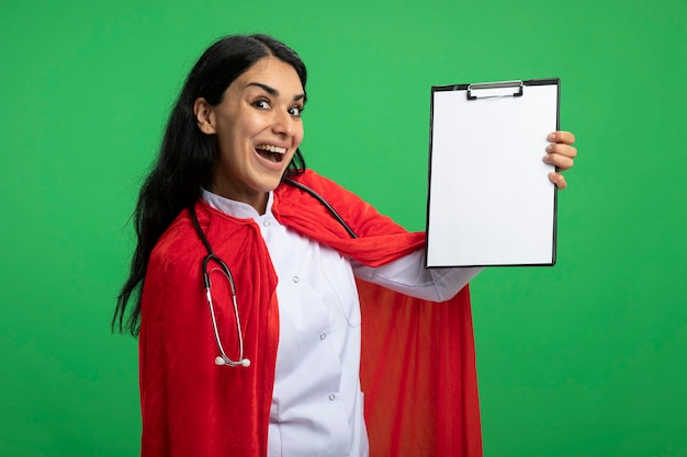 Alegre joven superhéroe vestida con bata médica con estetoscopio sosteniendo el portapapeles aislado en verde