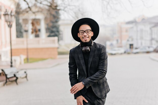 Alegre joven en traje oscuro y sombrero caminando por el parque en la mañana. Foto al aire libre del modelo masculino africano sonriente en ropa de moda
