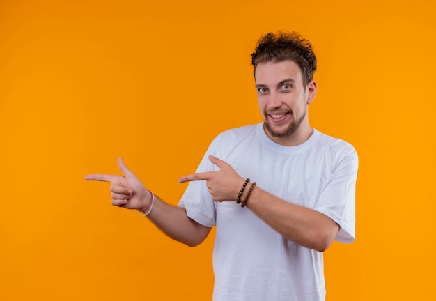 Alegre joven vestido con camiseta blanca apunta al lado con ambas manos en la pared naranja aislada