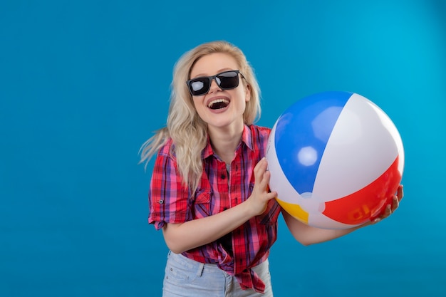 Alegre joven viajera con camisa roja en gafas sosteniendo una pelota inflable en la pared azul aislada