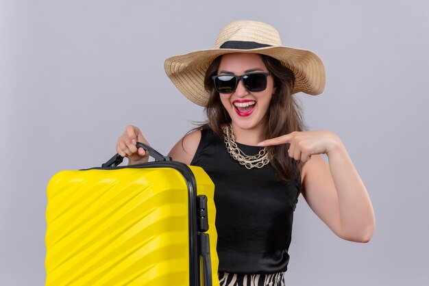 Alegre joven viajera vistiendo camiseta negra con sombrero en gafas sosteniendo maleta y apunta a la maleta en la pared blanca