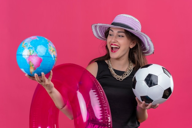 Alegre joven viajera vistiendo camiseta negra con sombrero sosteniendo círculo inflable y bola con globo en pared roja