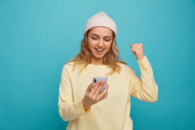 Alegre joven vistiendo gorro de invierno sosteniendo y mirando el teléfono móvil haciendo gesto de sí