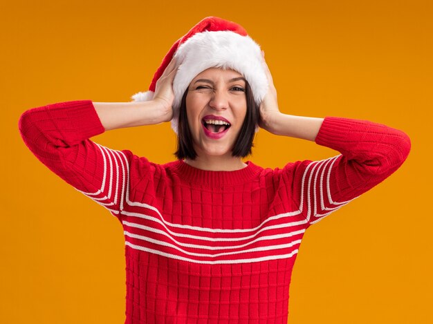 Foto gratuita alegre joven vistiendo gorro de papá noel mirando a la cámara guiñando un ojo manteniendo las manos en la cabeza aisladas sobre fondo naranja