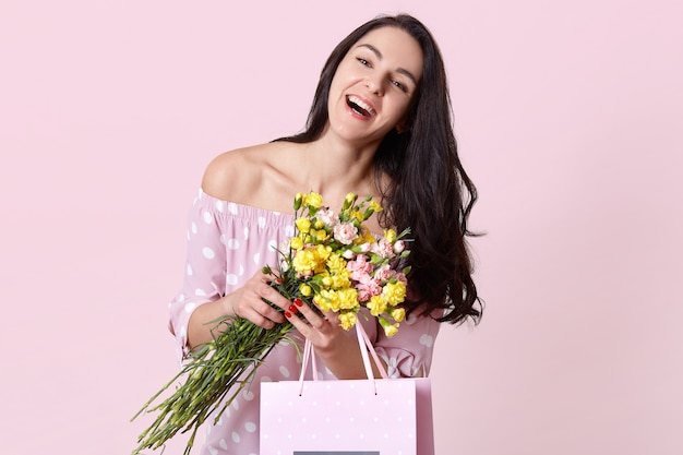 Alegre jovencita de cabello oscuro sonríe alegremente, vestida con ropa elegante, feliz de recibir el regalo en el cumpleaños, tiene un ramo de flores, modelos en rosa. Concepto de tiempo de primavera